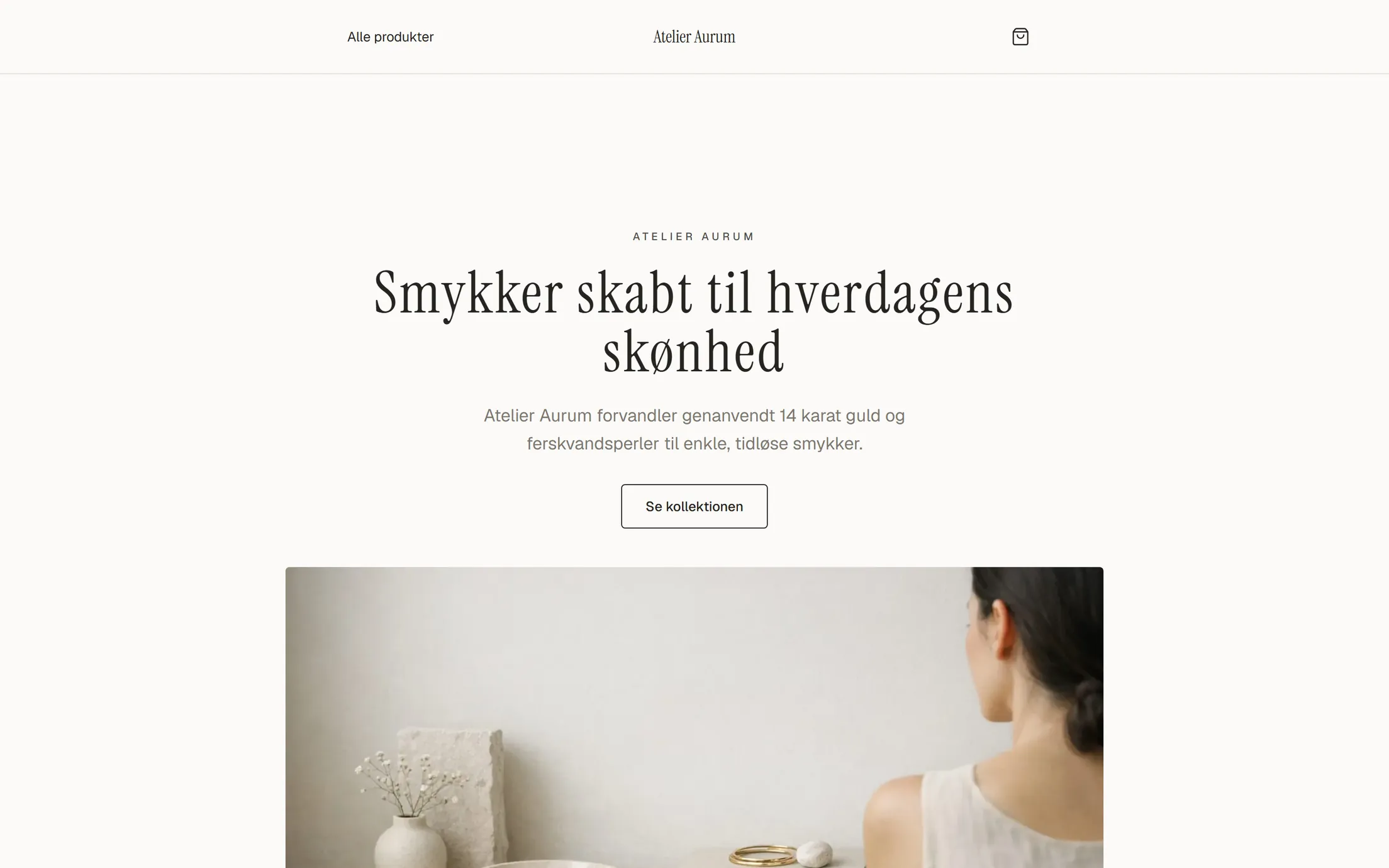 Demobutikken Atelier Aurum på en computerskærm: lys, varm hvid side med navnet centreret i en smal serif, overskriften 'Smykker skabt til hverdagens skønhed' centreret i serif, en tynd knap 'Se kollektionen', og under teksten et AI-lavet stilleben i sand og creme med guldringe.